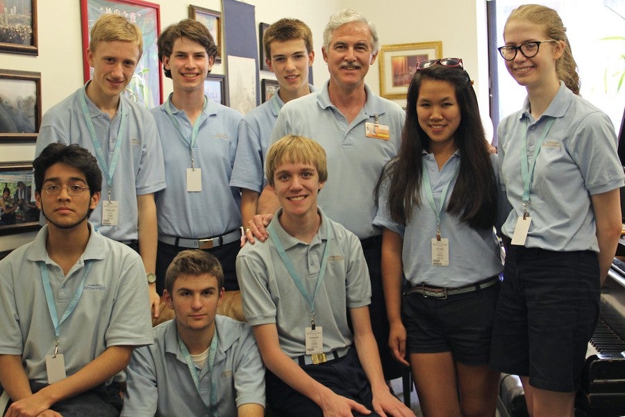 A male in his 60s with grey hair and a mustache poses stands with a group of eight high school-age students dressed in blue uniforms.